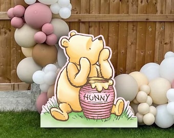 Pode incluir: Um recorte de Winnie the Pooh com um pote de mel que diz "Hunny". O recorte está na frente de uma cerca de madeira marrom e um arco de balões com balões brancos, bege e rosa.