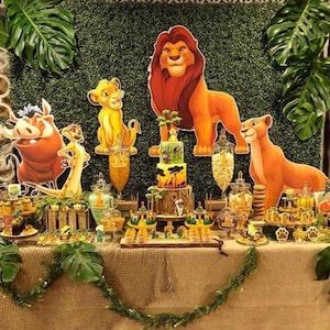 Könnte beinhalten: Eine Partydekoration mit Pappaufstellern eines Löwen, einer Löwin, eines Jungtieres, eines Warzenschweins und eines Erdmännchens. Eine dreistöckige Torte steht in der Mitte, umgeben von Gläsern mit Snacks, kleinen Leckereien und Monstera-Blättern.