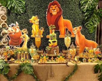 Puede incluir: Una decoración de fiesta con figuras de cartón de un león, una leona, un cachorro, un jabalí y una suricata. Un pastel de tres niveles se encuentra en el centro, rodeado de frascos de vidrio con bocadillos, dulces y hojas de monstera.