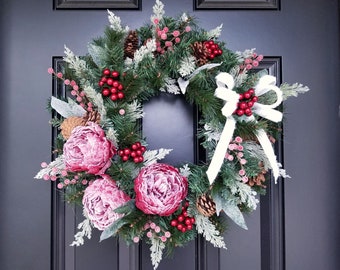 May include: A Christmas wreath with a white bow, red berries, pine branches, and pink peonies. The wreath is decorated with frosted pine cones and is hanging on a black door.
