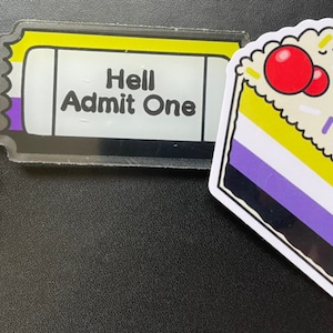 Może przedstawiać: Złoty brelok do kluczy z zawieszką w kształcie kwiatu i przezroczystym akrylowym biletem z nadrukiem "Hell Admit One". Bilet ma żółte, fioletowe i białe paski. Obok biletu znajduje się naklejka przedstawiająca kawałek ciasta z żółtymi, białymi, fioletowymi i czarnymi paskami. Ciasto ma na wierzchu czerwoną wiśnię i białe i czarne posypki.