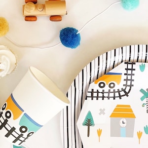 May include: A black and white striped paper plate with a white paper insert featuring a train track design with a yellow train, a gray building with a clock, and a green tree.