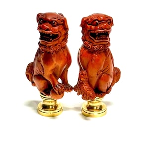 May include: Two carved wooden guardian lion statues with fierce expressions, reddish-brown in color. Each statue sits atop a gold-colored base. The lions have detailed manes and are holding a ball. Decorative home decor.