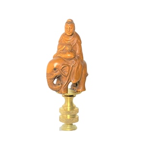 May include: A lamp finial featuring a carved wooden figure of a person seated on an elephant. The figure and elephant are a warm brown color, mounted on a brass base with a textured design. The finial is designed to be attached to a lamp.