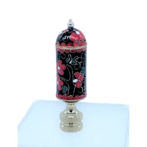 Puede incluir: Un remate de lámpara decorativo con un diseño floral negro, rojo y dorado. El remate tiene un cuerpo cilíndrico con una parte superior abovedada y una base de latón. El patrón floral incluye flores rojas y blancas sobre un fondo negro. El remate mide unos 10 cm de alto.