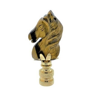 May include: A decorative lamp finial featuring a horse head design. The finial is made of a gold and black material, with a detailed horse head atop a gold-colored base. The finial is designed to be screwed onto a lamp.