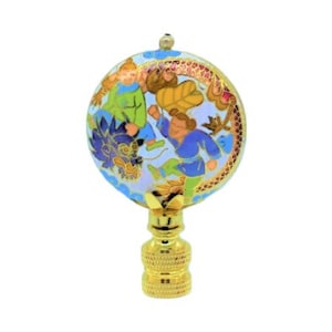 Puede incluir: Un remate de lámpara decorativo con base dorada y parte superior redonda esmaltada. La parte superior presenta una colorida ilustración de figuras en azul, verde y naranja, sobre un fondo azul claro con detalles florales. El remate mide unos 7.6 cm de alto.