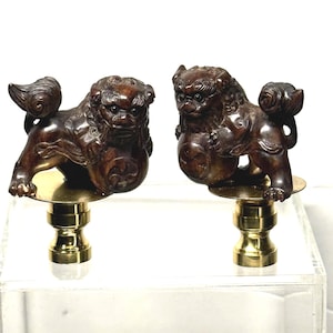 May include: Two brown, carved Foo Dog figurines. Each dog is posed with a ball and sits atop a gold-colored base. The figurines are displayed on a clear, rectangular surface.