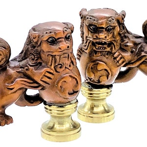 May include: Two intricately carved wooden guardian lion statues. The statues are brown with detailed features, mounted on brass bases. The lions are in a dynamic pose, with one appearing to be snarling. The statues are likely decorative items.