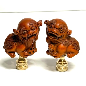 May include: Two carved wooden guardian lion figurines with detailed features, including open mouths and expressive eyes. The lions are mounted on brass bases. The wood has a warm, reddish-brown tone, and the overall style is traditional.
