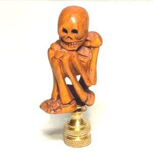 May include: A carved wooden skeleton figurine with a brass base. The skeleton is in a seated position, with its arms crossed and legs bent. The wood has a warm, orange-brown hue. The brass base is a shiny gold color.