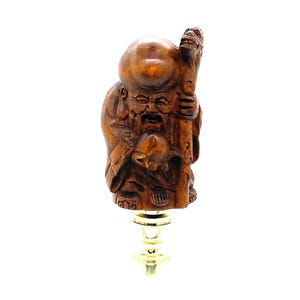 Peut inclure: Un bouchon de bouteille en bois représentant une figurine sculptée d'un vieil homme souriant avec une grosse tête et un bâton. La figurine est marron, avec une base en métal doré.