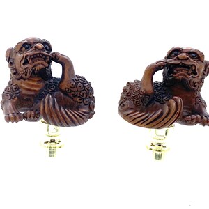May include: Two carved wooden figurines of mythical creatures. Each figure is brown with intricate details, featuring a lion-like body and a bird-like element. They are mounted on gold-colored bases, likely for a lamp or similar fixture.
