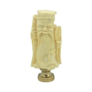 Peut inclure: Figurine ivoire d'un homme barbu souriant en tenue traditionnelle, tenant un éventail et un bâton. La figurine est montée sur une base en métal doré, probablement un embout de lampe. La sculpture présente des détails complexes.