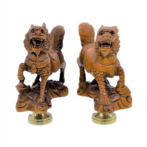 May include: Two intricately carved wooden statues of mythical creatures, possibly dragons or lions, with open mouths and detailed features. The statues are mounted on brass bases and rest on a carved wooden base. The wood has a warm, reddish-brown hue.
