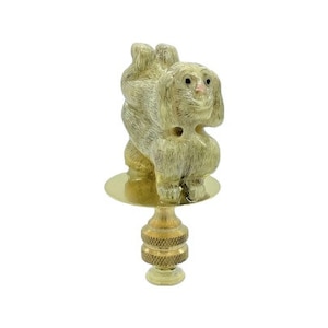 May include: A beige lamp finial featuring a carved dog figurine with a pink nose and black eyes. The dog sits atop a brass-colored base with a round disc and a decorative, textured stem.
