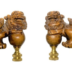 May include: Two wooden lamp finials carved in the shape of guardian lions. The lions are brown with detailed manes and fierce expressions. Each finial has a brass base with a threaded design, suitable for lamp assembly.