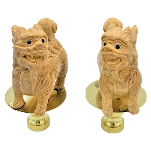 May include: Two carved wooden Foo Dog figurines. The dogs are light brown with detailed carvings, black eyes, and seated on gold-colored bases. The figurines are displayed against a white background.