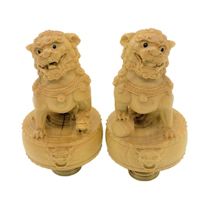 May include: Two wooden Foo Dog statues. Each statue is carved from light-colored wood and features a detailed lion-like figure with an open mouth, sitting atop a round base. The bases have decorative carvings and are approximately 3 inches in diameter.