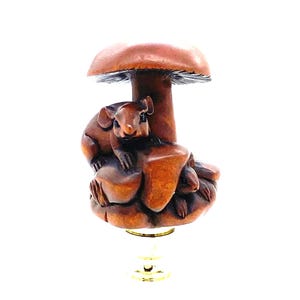 Puede incluir: Un remate de lámpara de madera con un hongo y dos ardillas. Las ardillas están talladas en la madera, con un sombrero de hongo detallado. El remate tiene una base dorada y está sobre un fondo blanco.
