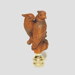 May include: A wooden lamp finial featuring two carved birds perched together. The birds are a warm brown color, with detailed feather carvings. The finial has a brass base with a threaded design.