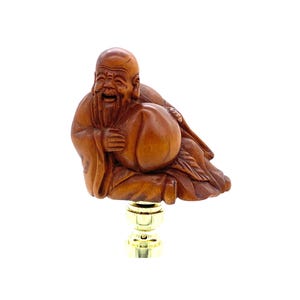 Peut inclure: Un embout de lampe en bois représentant une figurine sculptée d'un homme souriant avec une longue barbe, tenant une grosse pêche. La figurine est marron et repose sur une base dorée.