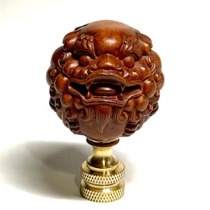 May include: A detailed, hand-carved wooden lamp finial featuring a mythical creature's head. The finial is a rich brown color, with intricate details and a brass base. The design is reminiscent of traditional Asian art.