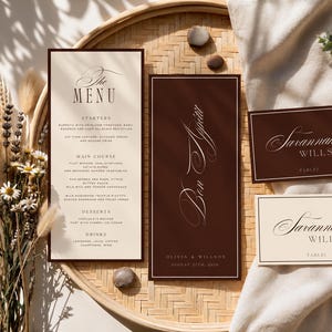May include: A set of wedding stationery including a tall cream menu card with black text, a tall dark brown card with white text reading Bon Appétit, and two rectangular place cards with the name Savannah Willson in dark brown and cream.