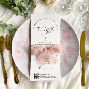 Puede incluir: Un lugar de boda con una tarjeta blanca con la frase "THANK YOU" y una cinta rosa. La tarjeta está sobre un plato con estampado rosa y blanco, con cubiertos dorados.