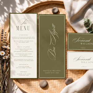 Può includere: Un set di cancelleria per matrimoni composto da un menu alto con sezioni color crema e verde oliva, una carta alta abbinata verde oliva con il testo Bon Appétit e due segnaposto rettangolari con il nome Savannah Willson in carattere serif.