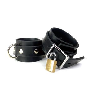 May include: Black leather restraints with a silver buckle and a gold padlock. The padlock has the text "BASI" on it.