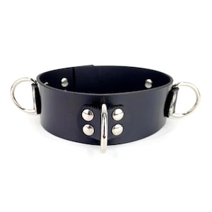 Op de afbeelding: Een zwarte halsband met een gladde, brede band en drie zilverkleurige metalen ringen. De middelste ring is bevestigd aan een rechthoekig leren lipje met vier zilveren klinknagels, terwijl er aan de zijkanten nog twee ringen zitten.