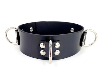 May include: A black collar featuring a smooth, wide band with three silver-toned metal rings attached. The central ring is secured by a rectangular leather tab with four silver rivets, while two additional rings are positioned on the sides.