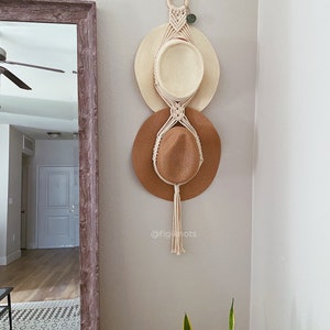 May include: A macrame hat rack with three straw hats hanging on it. The rack is made of white cord and has a wooden ring at the top. The hats are in different shades of brown and beige. The rack is hanging on a white wall. The text "@figiknots" is visible below the hats.