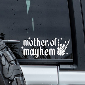May include: White vinyl decal sticker with the text "mother of mayhem" in a gothic font and a skeleton hand making the rock and roll hand gesture. The sticker is on the back window of a black car.