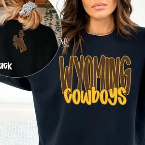 May include: A navy blue sweatshirt with the words "WYOMING COWBOYS" in a brown and yellow font. The back of the sweatshirt features a brown graphic of a cowboy riding a horse. The sweatshirt is a crew neck style.