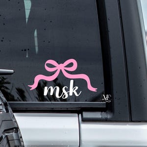 May include: Pink bow with a white "msk" written underneath. The bow and text are on a black car window.