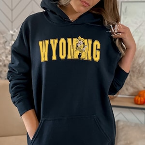 May include: Black hoodie with a yellow graphic of a cowboy wearing a hat and holding a gun. The text "WYOMING" is printed in yellow above the graphic.