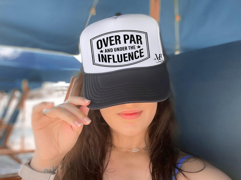 May include: A white and black trucker hat with the text "Over Par and Under the Influence" in a black rectangle. The hat has a black mesh back.