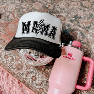 May include: A white trucker hat with a black bill and the word "MAMA" in black letters with a checkered lightning bolt design. The hat is sitting on a silver disco ball.
