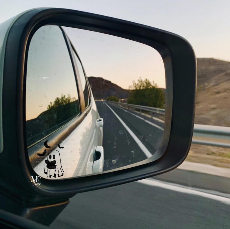 May include: A car's side mirror reflecting a road scene at dusk. A white car is visible, with a ghost and bat sticker on the mirror. The road stretches into the distance, flanked by a guardrail and a mountain.