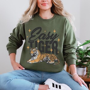 K&ouml;nnte beinhalten: Ein gr&uuml;ner Pullover mit dem Text "Easy Tiger" und einem Comic-Tiger, der auf der Vorderseite aufgedruckt ist. Der Tiger ist orange und schwarz mit wei&szlig;em Hintergrund.