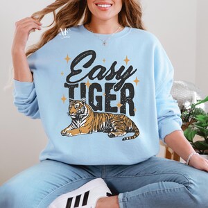 K&ouml;nnte beinhalten: Ein hellblauer Pullover mit dem Text "Easy Tiger" in schwarzen Buchstaben und einer stilisierten Tigerillustration. Der Tiger ist orange und schwarz mit goldenen Sternen um den Text.