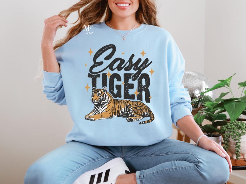 K&ouml;nnte beinhalten: Ein hellblauer Pullover mit dem Text "Easy Tiger" in schwarzen Buchstaben und einer stilisierten Tigerillustration. Der Tiger ist orange und schwarz mit goldenen Sternen um den Text.