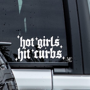 May include: A white vinyl sticker with the text "hot girls hit curbs" in a gothic font. The sticker is on the rear window of a black car.