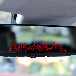 May include: A black car rearview mirror with a red flame decal across the bottom. The flames are stylized and add a unique, eye-catching detail to the vehicle's interior. The mirror reflects a blurred outdoor scene.