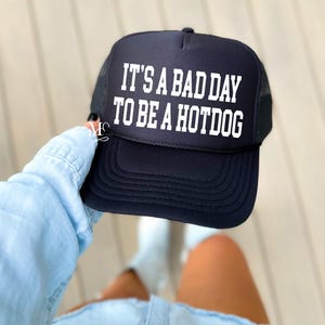 May include: Black trucker hat with a white mesh back. The hat has white text that reads "It's a bad day to be a hotdog."