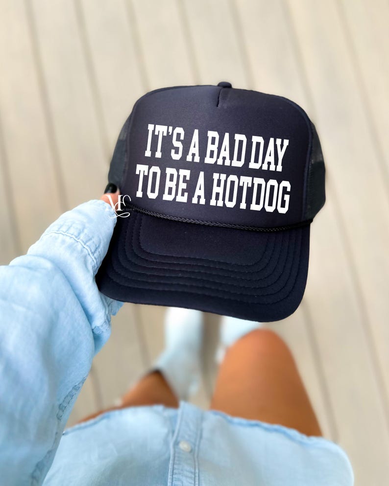 May include: Black trucker hat with a white mesh back. The hat has white text that reads "It's a bad day to be a hotdog."
