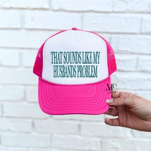 May include: A pink and white trucker hat with the text "THAT SOUNDS LIKE MY HUSBANDS PROBLEM" in green. The hat has a mesh back and a curved brim. The hat is held by a hand with rings.