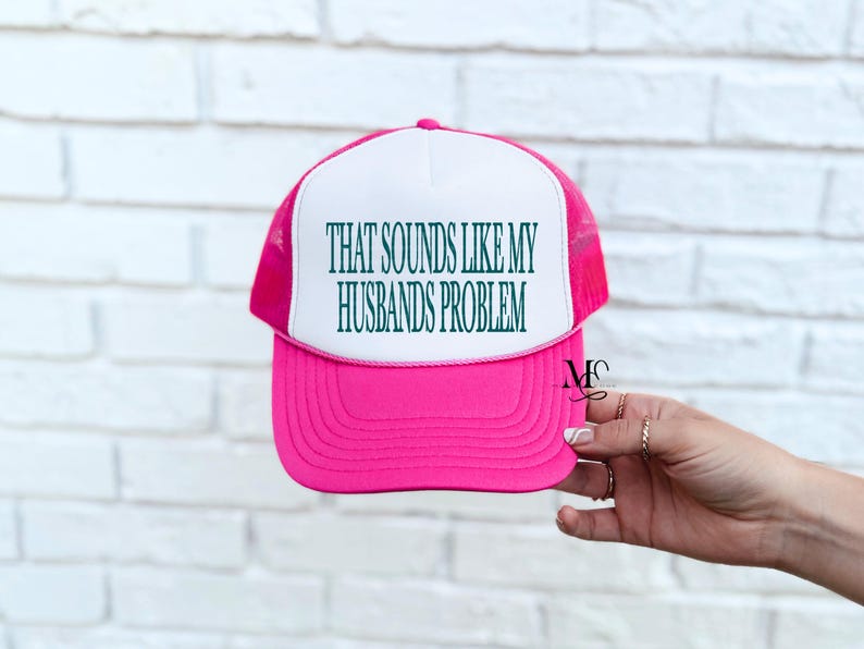 May include: A pink and white trucker hat with the text "THAT SOUNDS LIKE MY HUSBANDS PROBLEM" in green. The hat has a mesh back and a curved brim. The hat is held by a hand with rings.